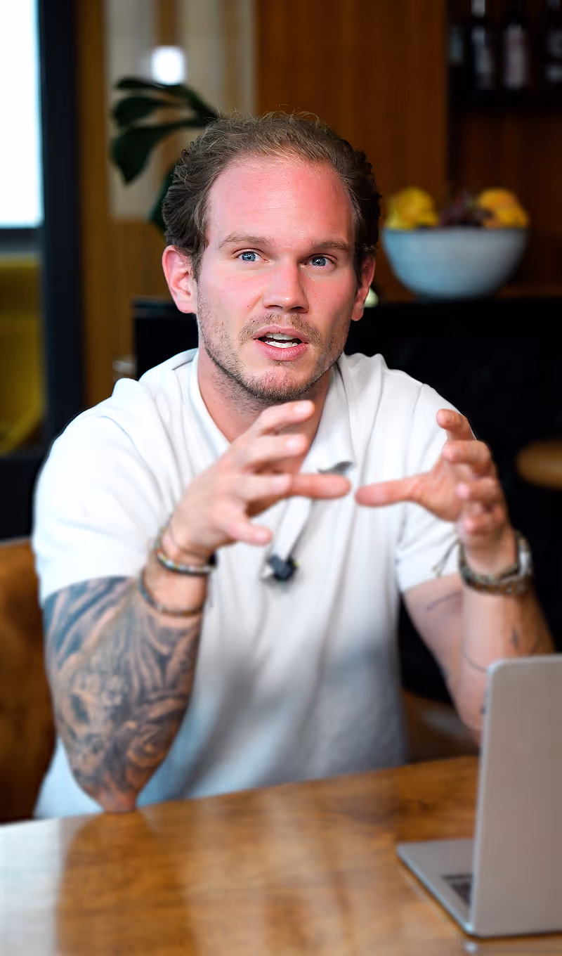 Junger Mann mit Tattoos und weißem Hemd erklärt etwas mit beiden Händen über einem Holztisch mit Laptop.
