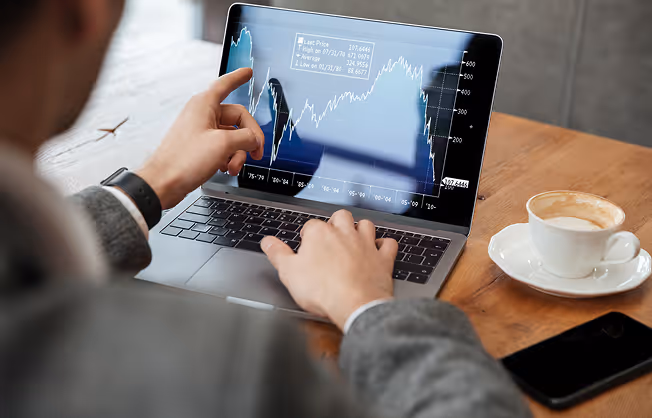 Person analysiert Aktienkursdiagramm auf Laptop, neben einer Tasse Kaffee und einem Smartphone.
