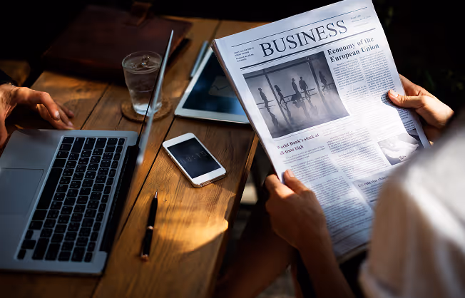Person reading a business newspaper by a wooden table with laptop, phone, pen, and glass of water.