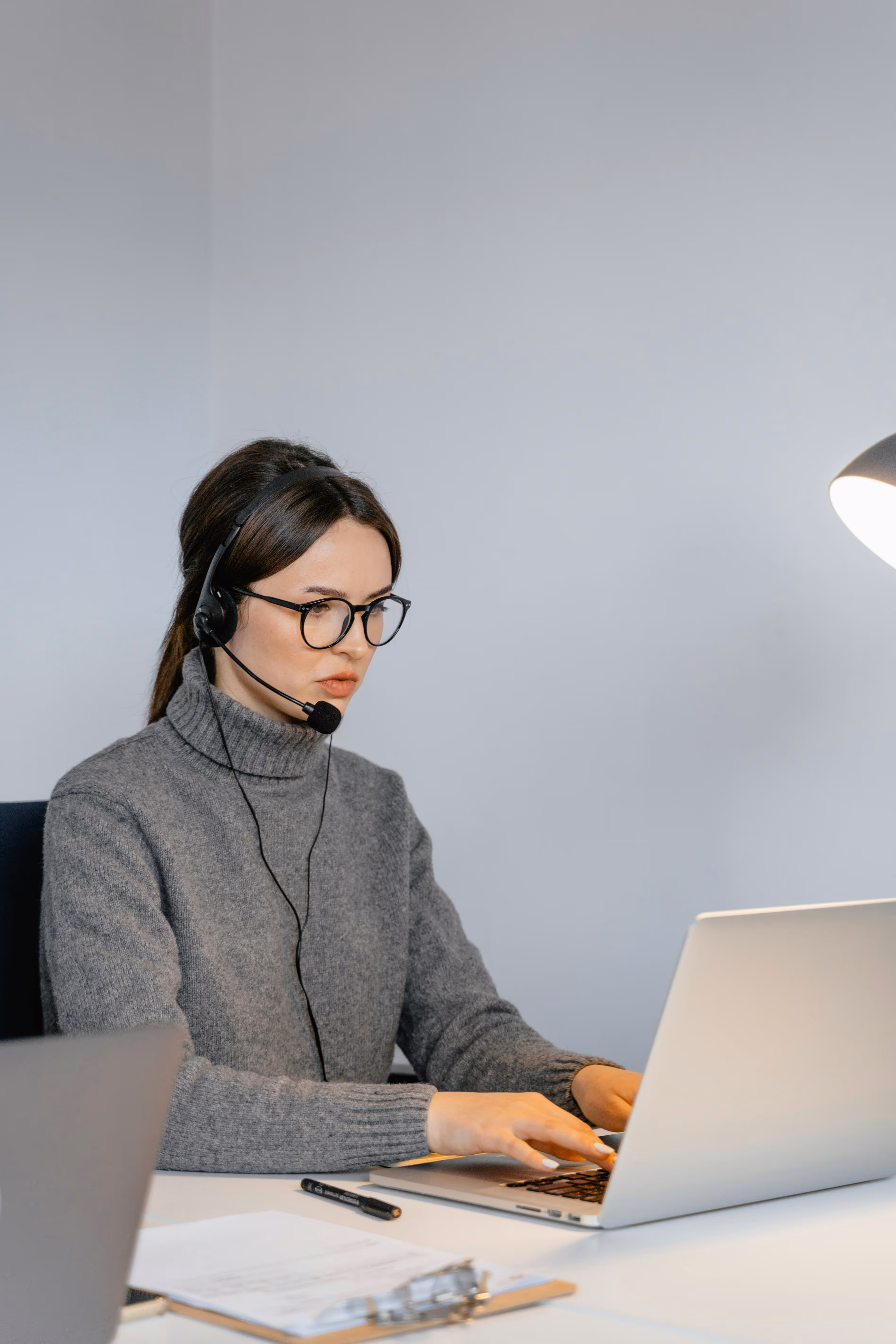 Frau mit Headset und Brille arbeitet konzentriert an Laptop in Büro mit grauem Pullover.