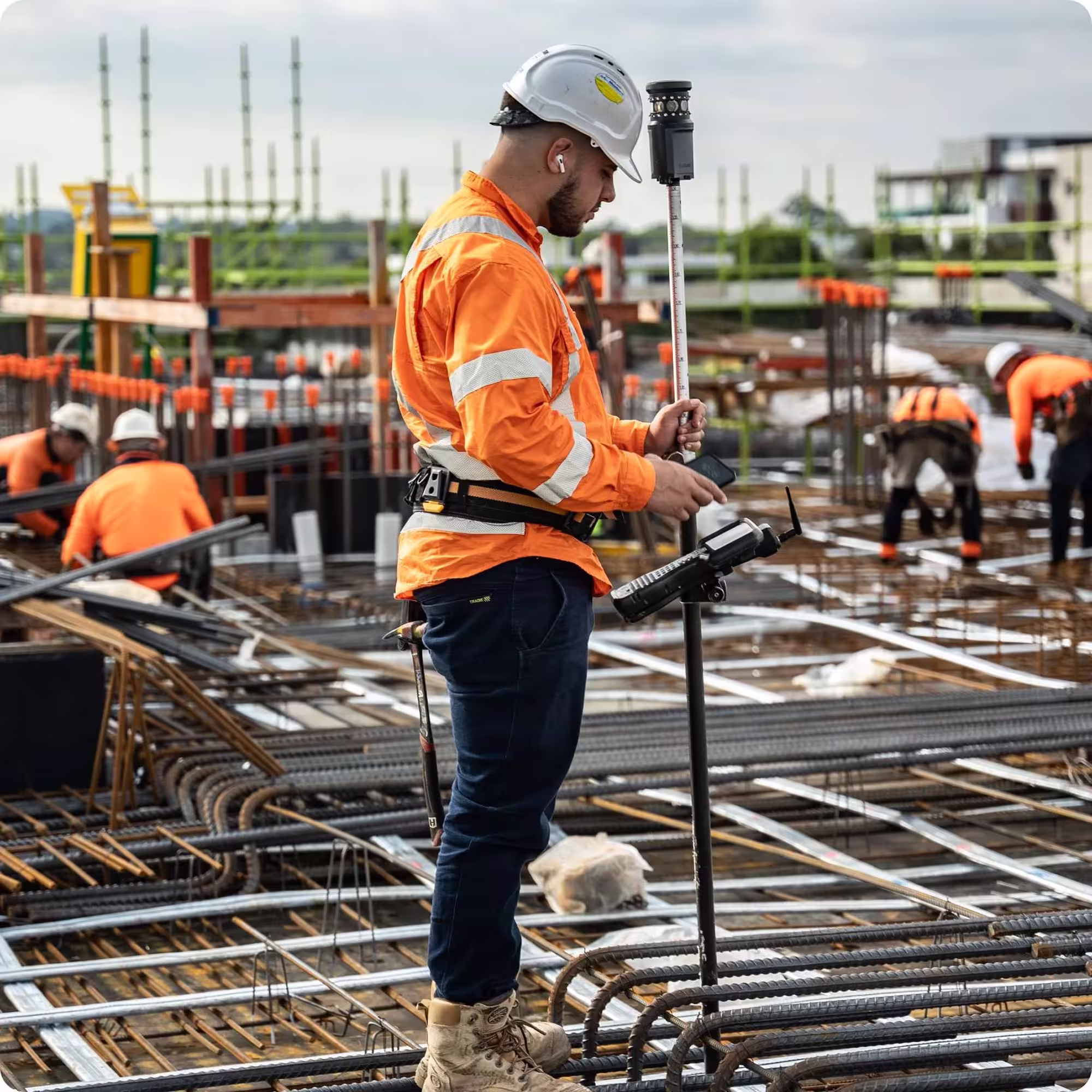 Construction worker in orange safety jacket and white helmet using surveying equipment on a building site with steel rebar and other workers in the background.