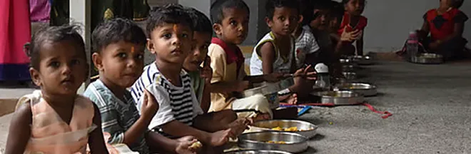 A group of young children sitting in a row on the floor eating food from metal plates.