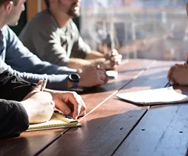 People sitting at a wooden table writing notes with pens, with sunlight coming through a window.