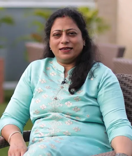 Smiling woman in a light blue embroidered traditional outfit sitting outdoors on a wicker chair.