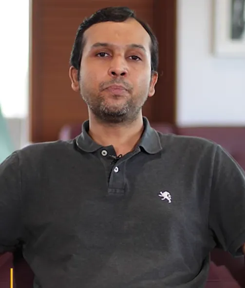 Man wearing a dark grey polo shirt sitting indoors with a neutral expression.