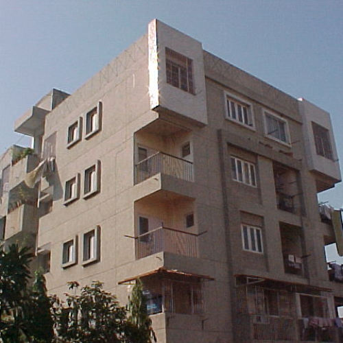 Four-story beige residential building with multiple windows, balconies, and plants at the base under a clear sky.