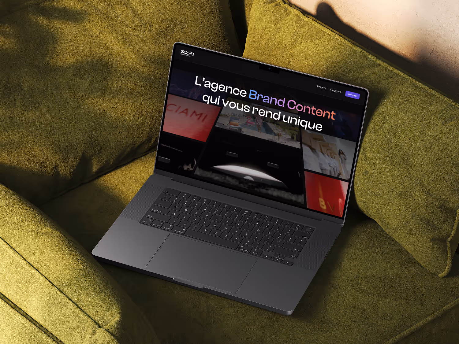 Open laptop on green couch showing Scob homepage with French text 'L'agence Brand Content qui vous rend unique'.