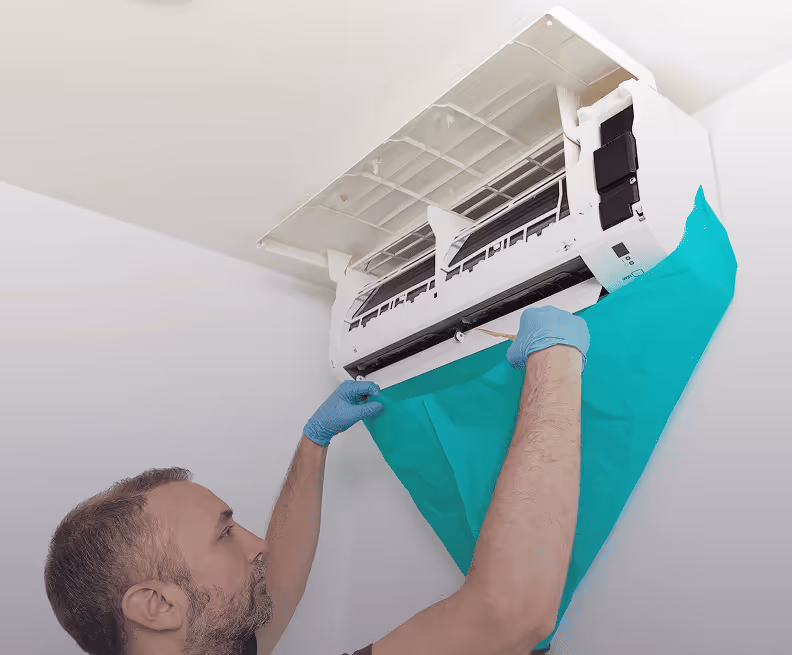 Un homme portant des gants bleus installe une couverture verte sous un climatiseur mural blanc ouvert pour l'entretien.