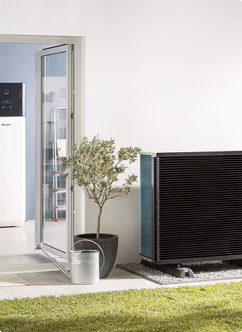 Système de pompe à chaleur noir installé à l'extérieur près d'une porte vitrée ouverte avec un petit arbre en pot et un arrosoir en métal à côté.