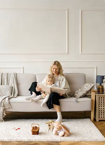 Une femme souriante lit un livre avec une petite fille assise sur ses genoux sur un canapé dans un salon clair.