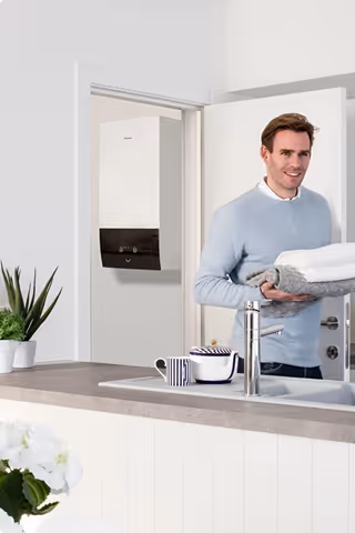 Un homme souriant portant des serviettes dans une cuisine moderne avec un chauffe-eau mural en arrière-plan.