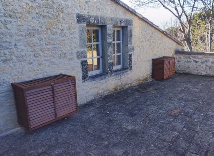 Façade en pierre avec une fenêtre à cadre blanc et deux boîtiers en bois brun posés sur un sol pavé extérieur.