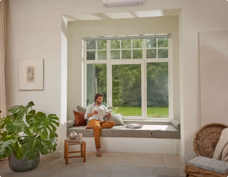 Un homme assis sur un coussin à une fenêtre avec vue sur un jardin verdoyant, entouré de plantes d'intérieur et de décorations naturelles.