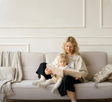 Une femme et une petite fille lisent un livre ensemble assises sur un canapé beige dans un salon lumineux.
