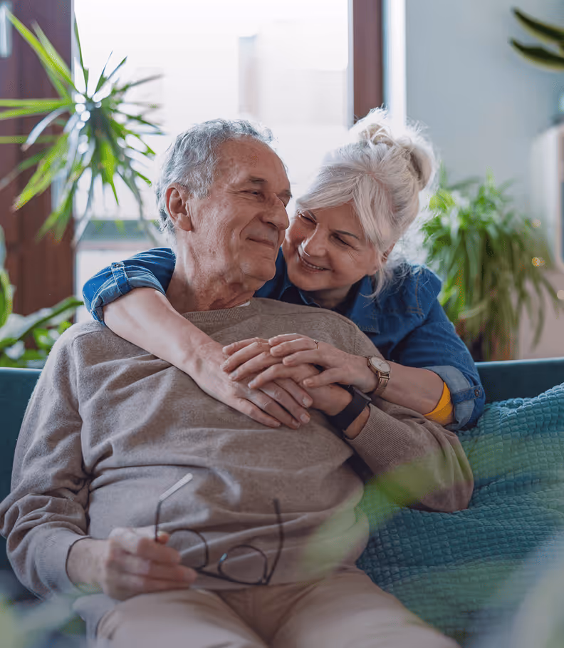 Un couple âgé souriant s'enlace sur un canapé entouré de plantes d'intérieur.