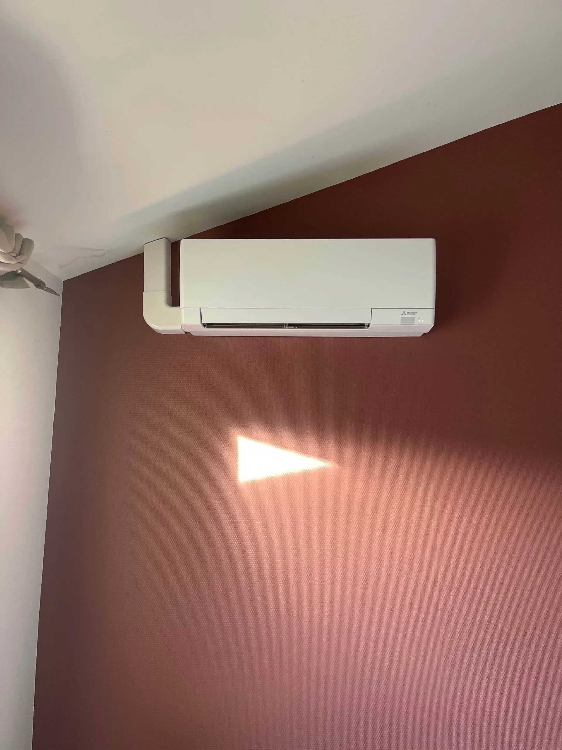 Climatiseur mural blanc installé sur un mur rouge avec une lumière en forme de triangle projetée dessous.