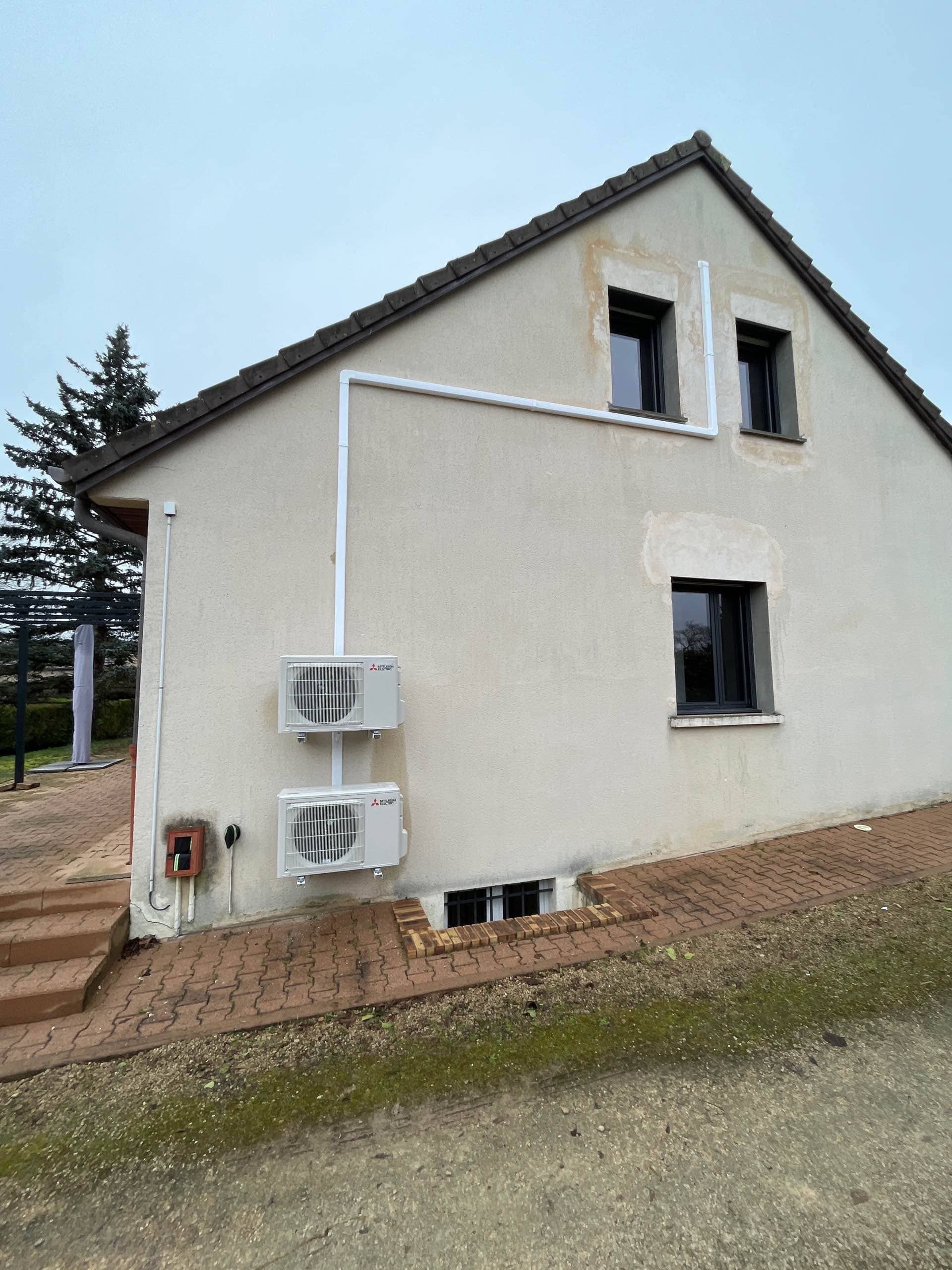 Façade latérale d'une maison beige avec deux unités extérieures de climatisation et trois fenêtres.
