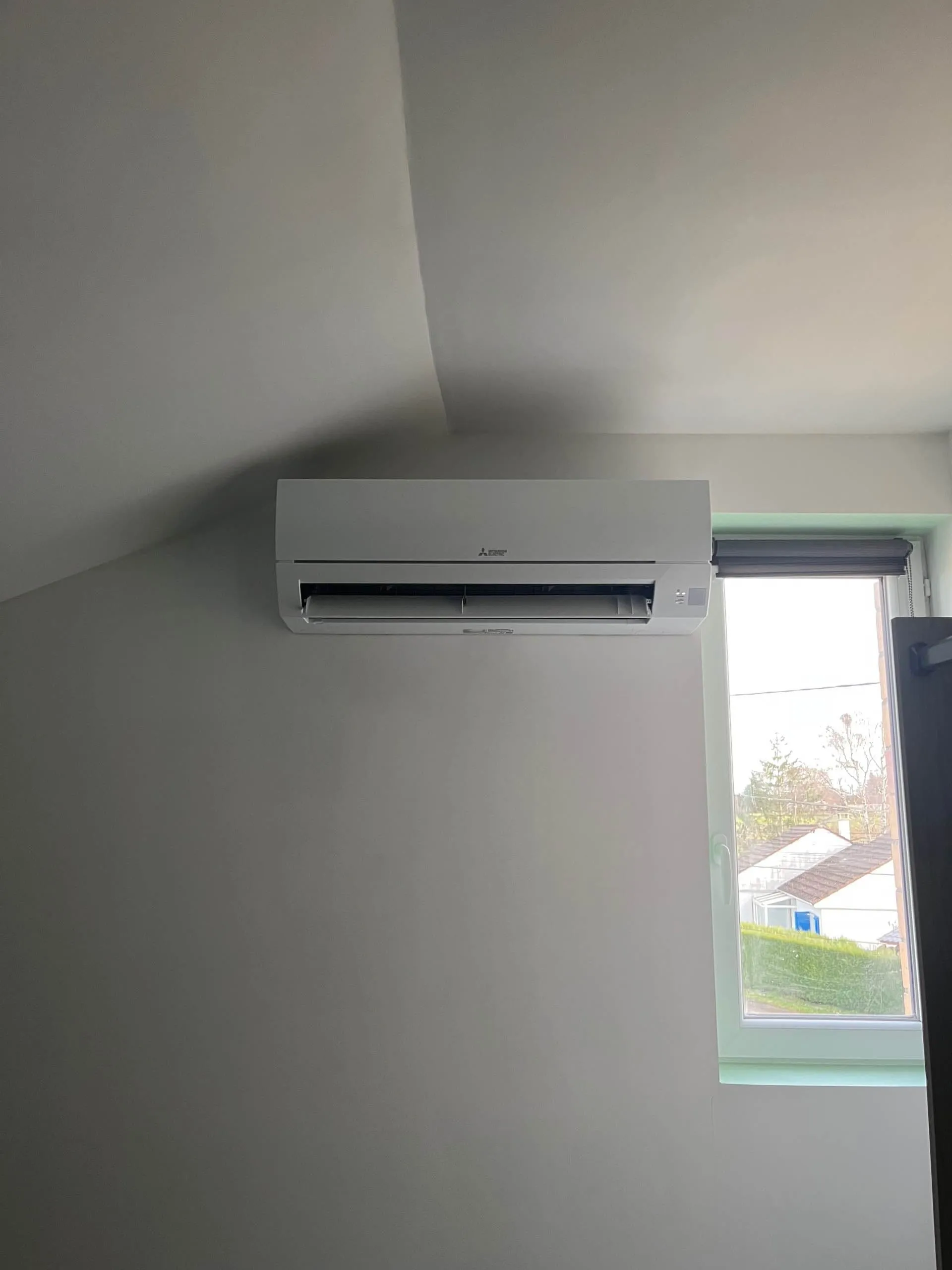 Unité intérieure de climatisation Mitsubishi installée au mur près d'une fenêtre dans une pièce aux murs clairs.