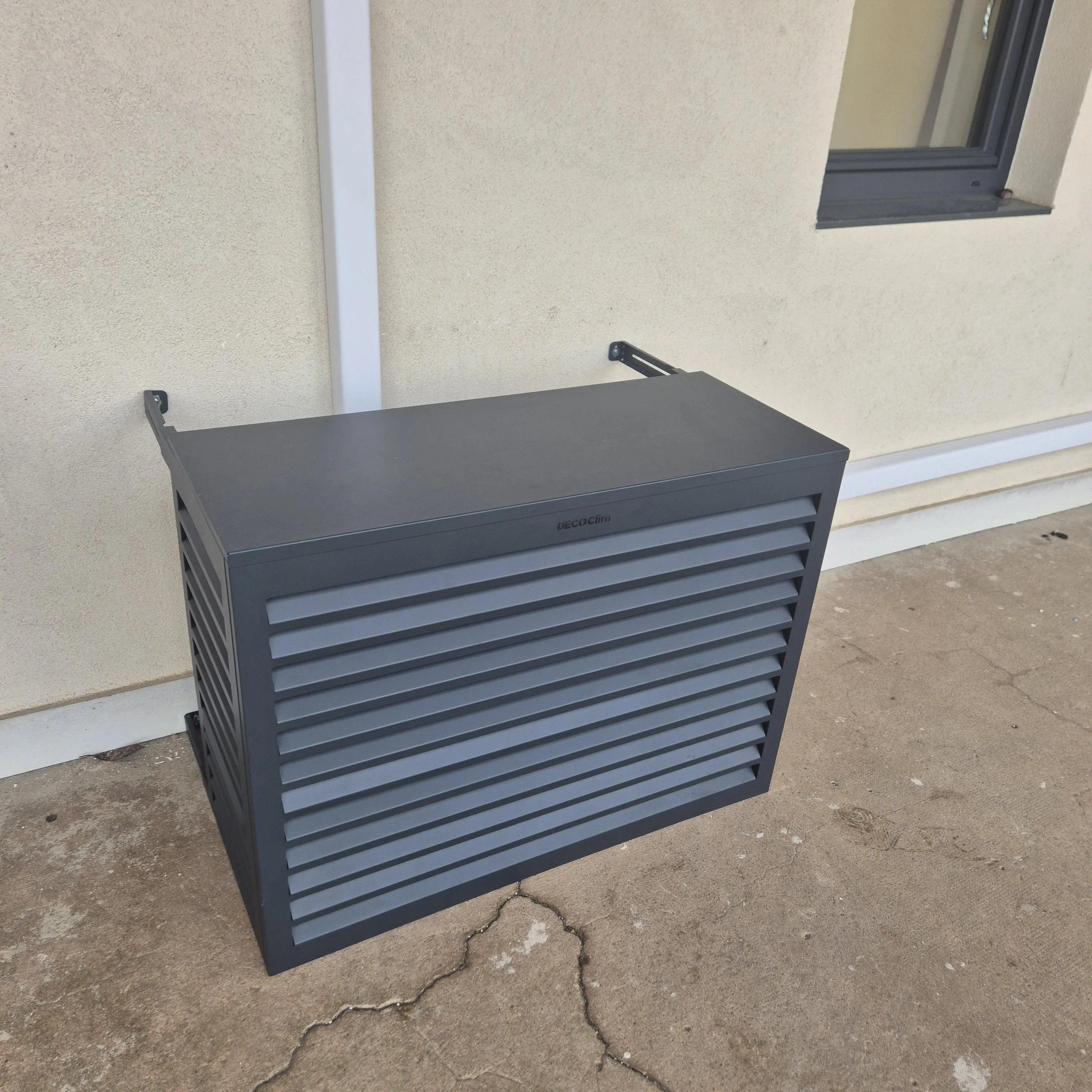 Protection en métal gris avec lattes horizontales installée devant une unité de climatisation extérieure contre un mur beige.