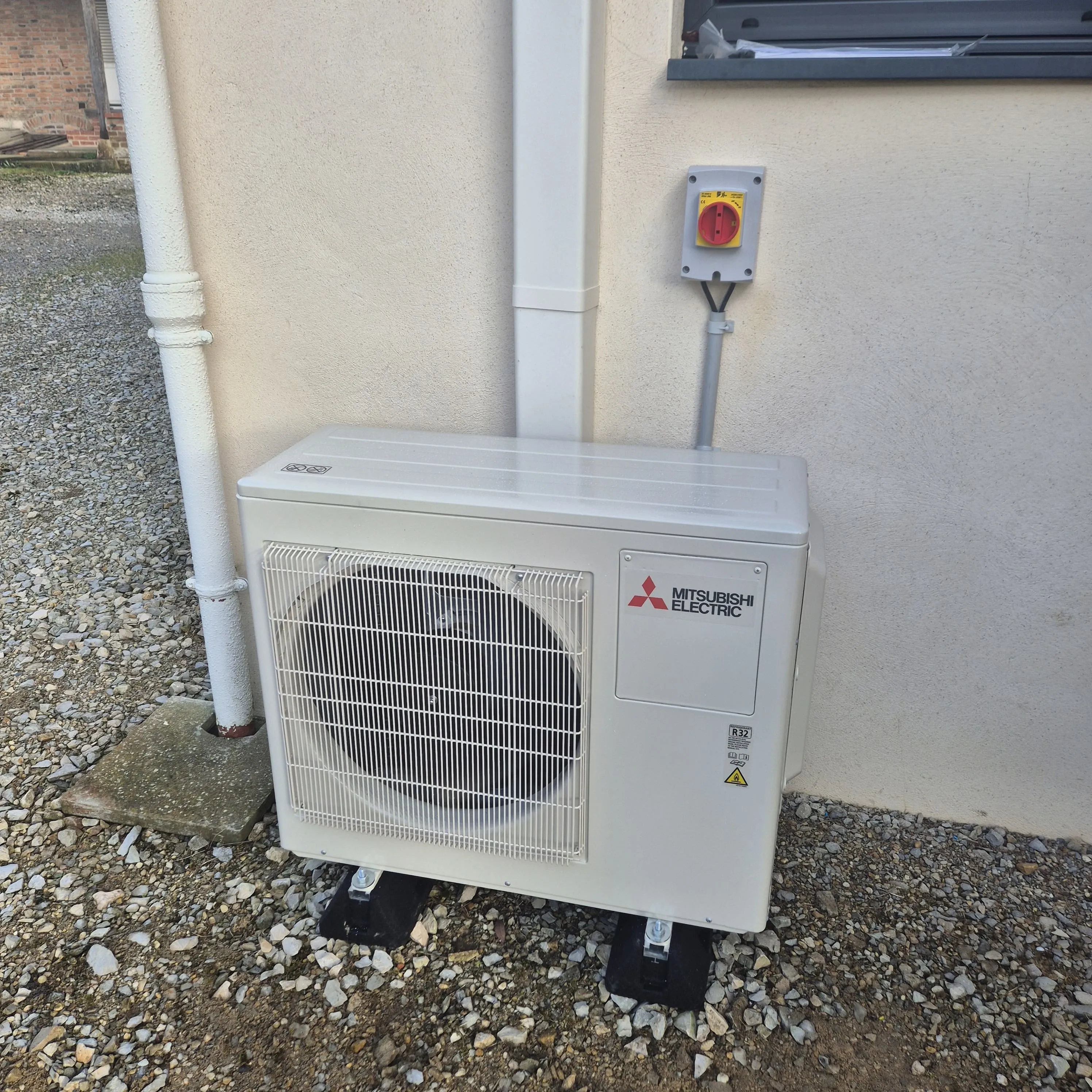 Unité extérieure d'un système de climatisation Mitsubishi Electric installée contre un mur, posée sur des supports noirs sur un sol gravillonné.