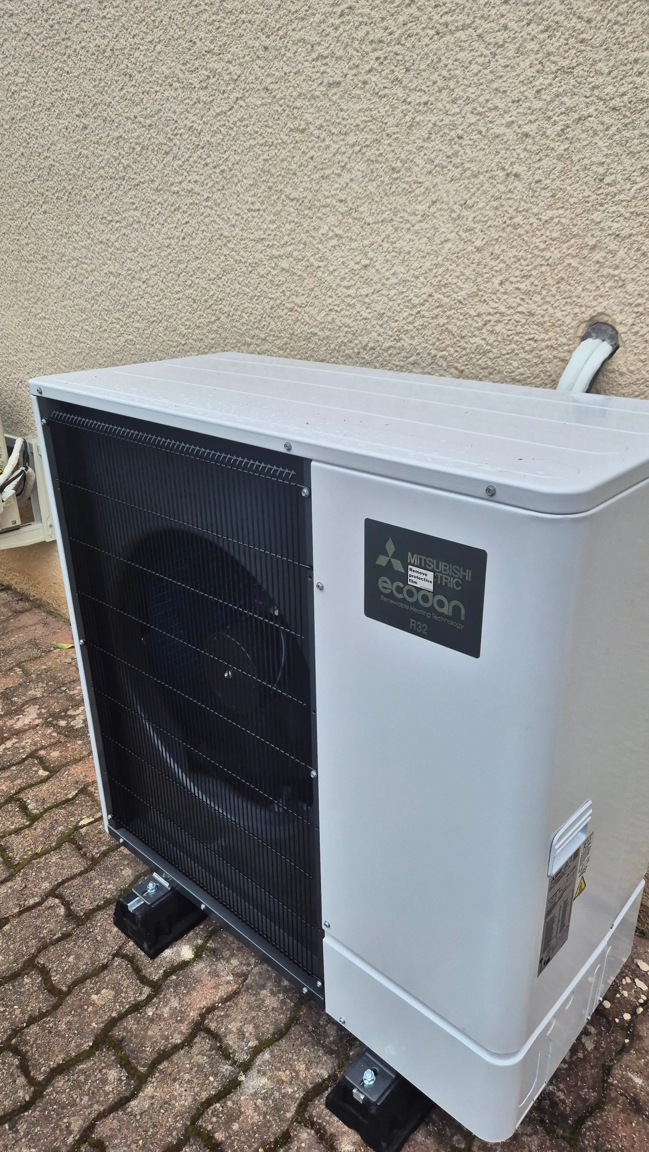 Unité extérieure de pompe à chaleur Mitsubishi Electric Ecoan installée sur un sol pavé à côté d’un mur beige.