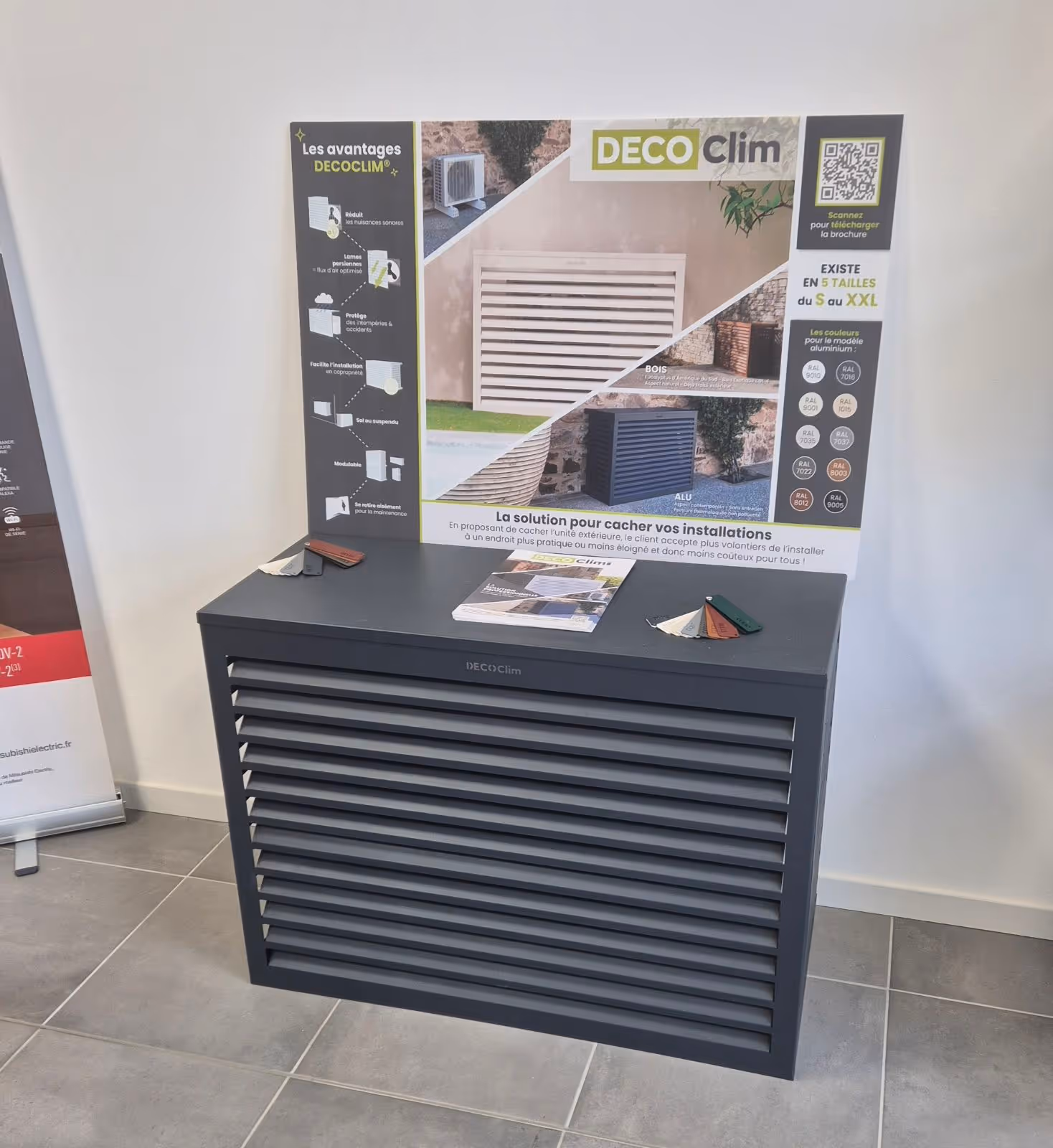 Un cache pour unité extérieure DECOclim gris avec brochures et échantillons de couleurs sur le dessus, placé devant une affiche explicative sur les avantages et options de taille et couleur.
