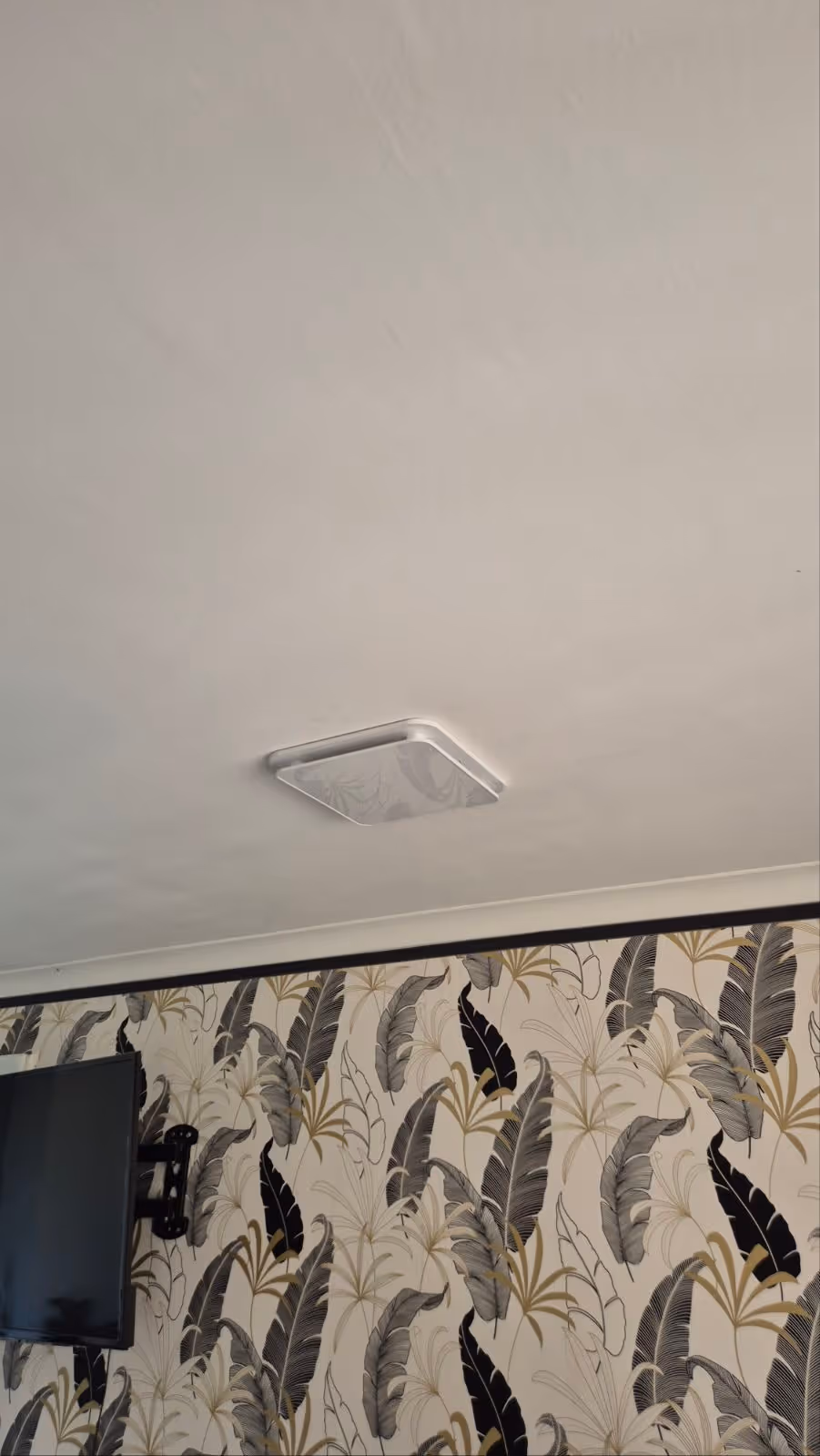 Climatiseur encastré au plafond d'une pièce avec papier peint à motifs de grandes feuilles grises, noires et beiges et un téléviseur mural.