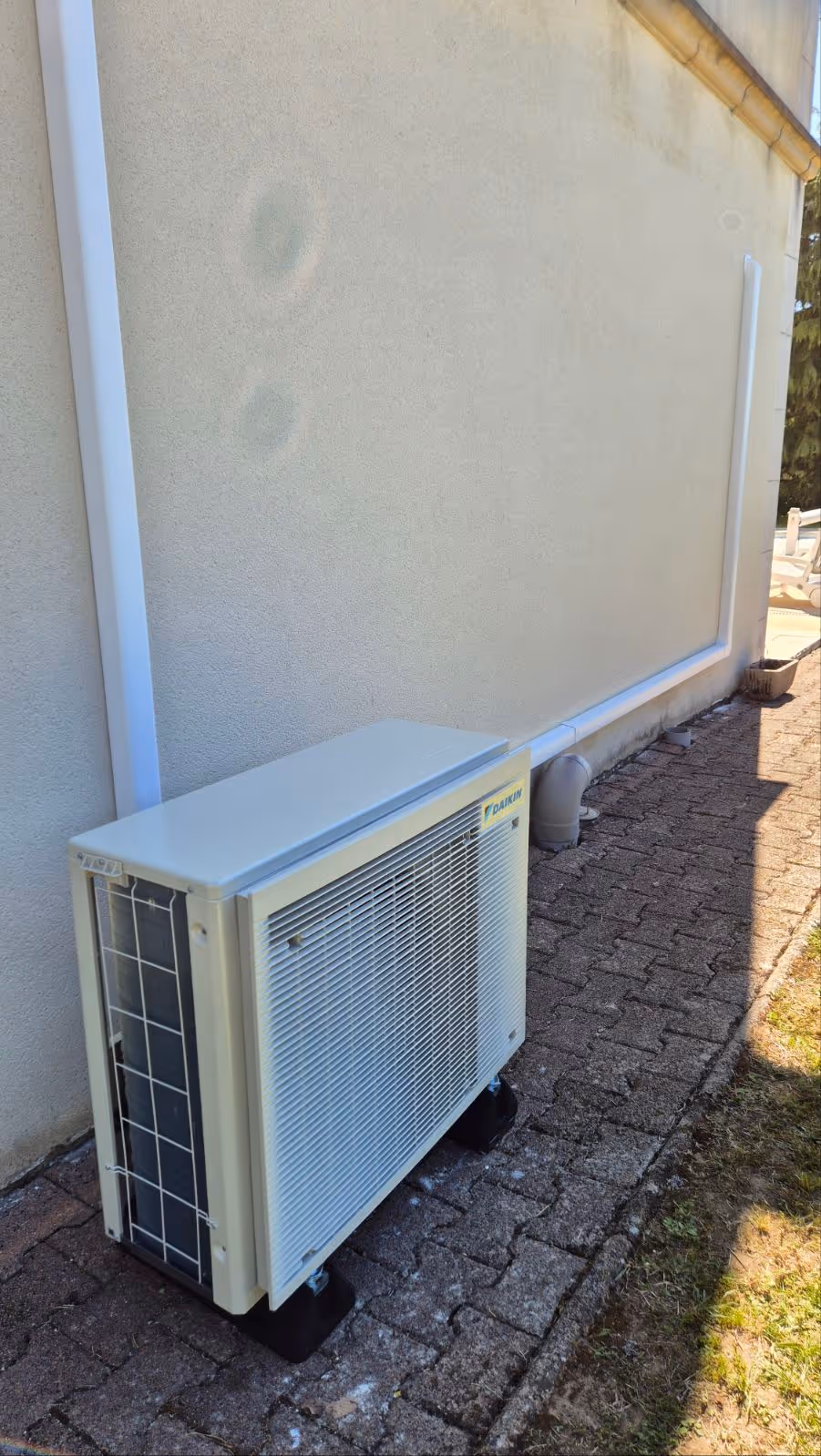 Unité extérieure de climatisation Daikin installée sur un sol en pavés contre un mur beige avec conduits blancs le long du mur.