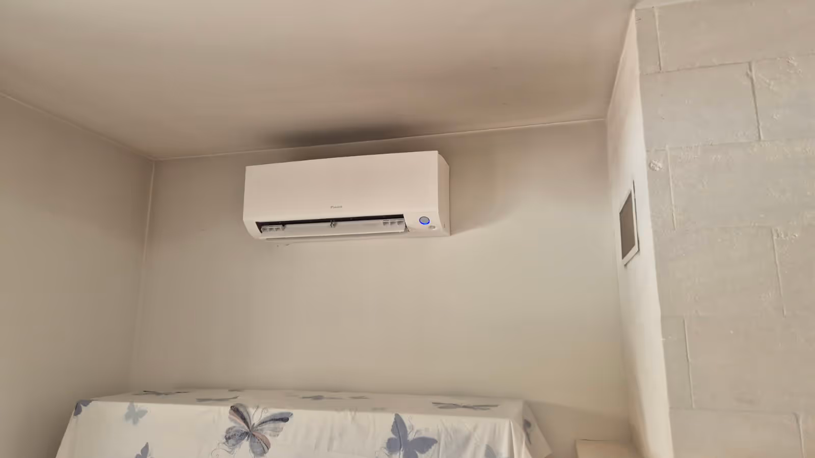 Climatiseur mural blanc Daikin installé dans un coin de pièce avec un meuble couvert d'une housse à motifs papillons.