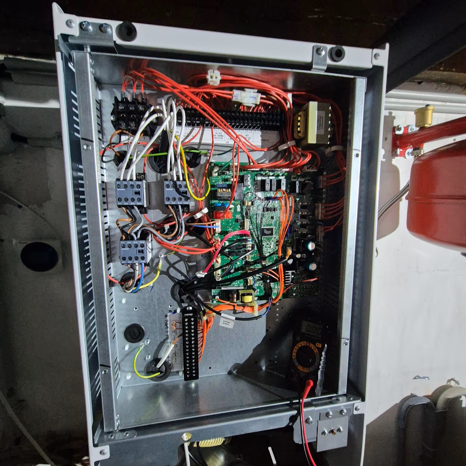 Coffret électrique ouvert montrant des circuits imprimés, des relais, de nombreux fils rouges, blancs et noirs connectés, et un multimètre noir posé à l'intérieur.