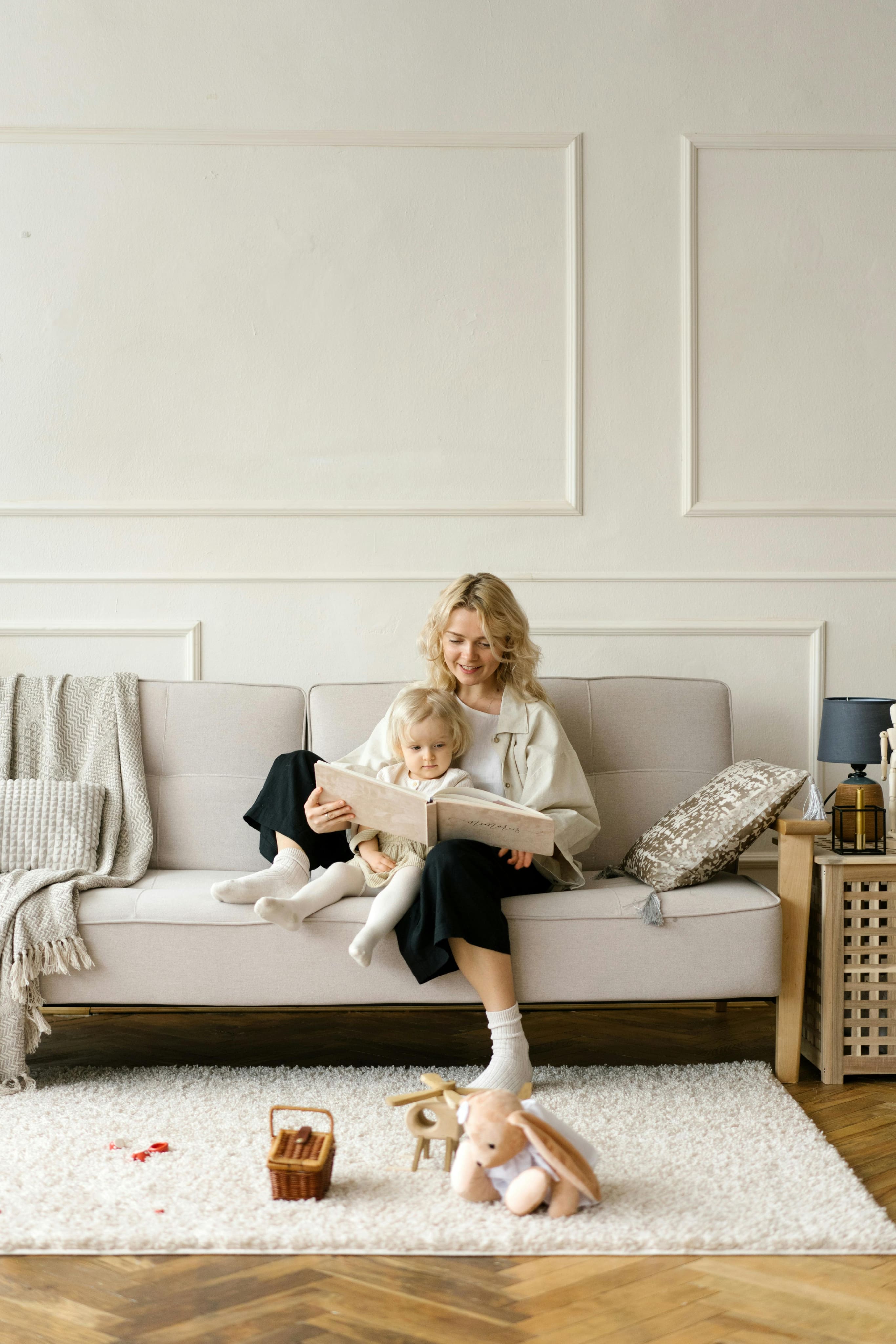 Une femme souriante lit un livre avec une petite fille assise sur un canapé beige dans un salon lumineux et décoré avec des coussins, une couverture, des plantes et des jouets sur un tapis blanc.