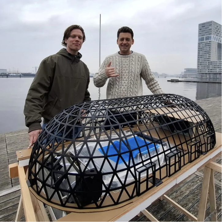 Twee mannen poseren bij een zwart geometrisch model van een gebouw aan het water.
