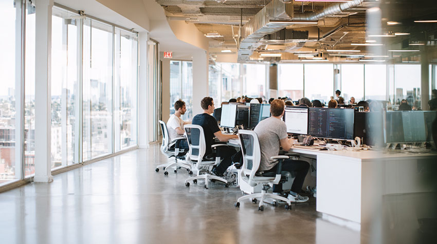 A candid, documentary-style photograph of a bustling tech startup office. Diverse young professionals in casual clothes (hoodies, jeans) are intensely focused, working on laptops displaying lines of code and data interfaces at long communal reclaimed wood tables.