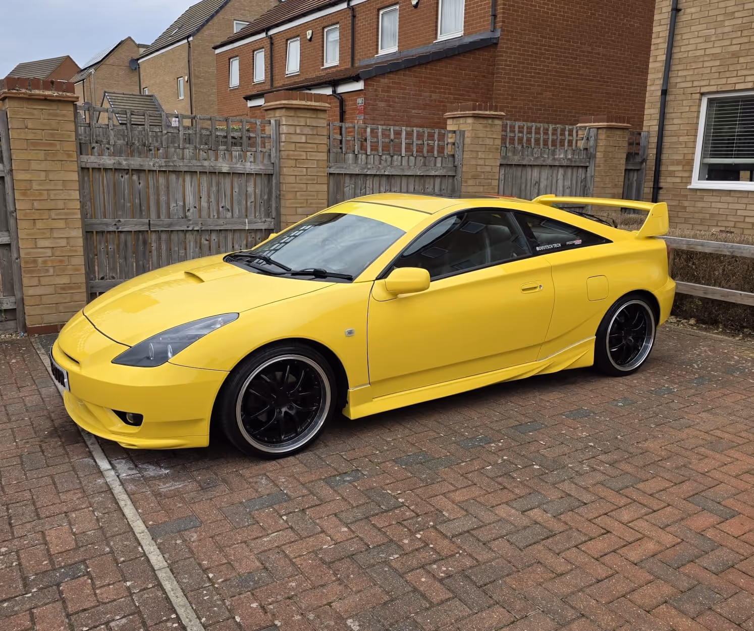 Yellow sports coupe with black rims parked on a brick driveway in front of a wooden fence and brick houses.