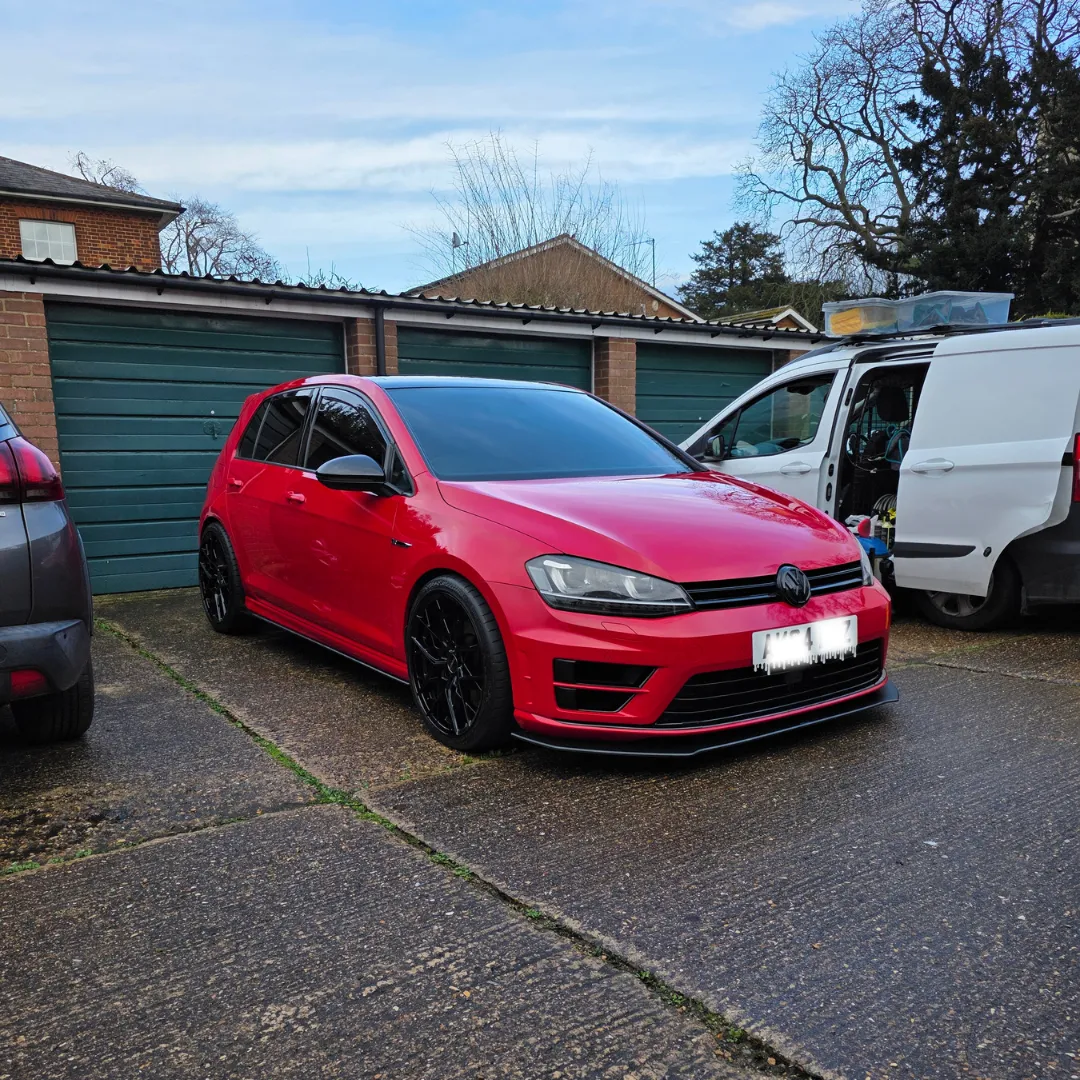 Red Volkswagen hatchback with black rims parked on driveway in front of green garage doors, next to a white van.