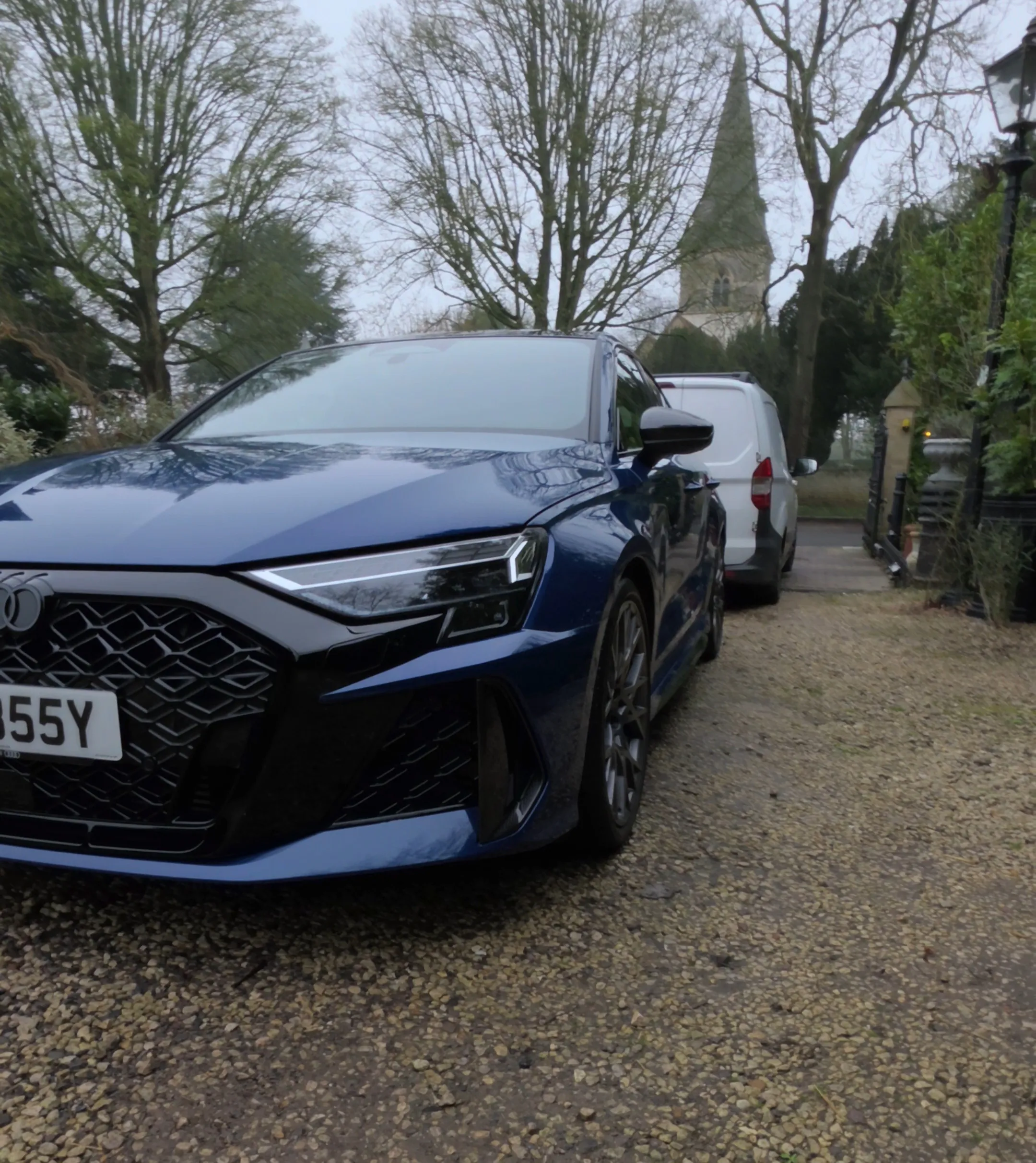Blue Audi car parked on a gravel driveway in front of a white van with trees and a church steeple in the background.