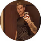 Junger Mann mit Tattoo am linken Arm macht Selfie vor einem Spiegelschrank.