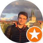 Porträt eines Mannes mit dunklen Haaren und schwarzer Jacke vor einem städtischen Hintergrund mit historischer Architektur und bewölktem Himmel.