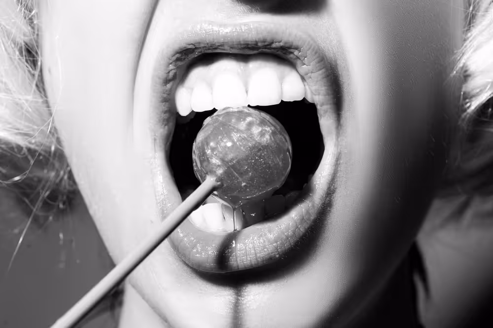 Close-up of an open mouth with a round lollipop resting on the tongue, showing healthy white teeth.
