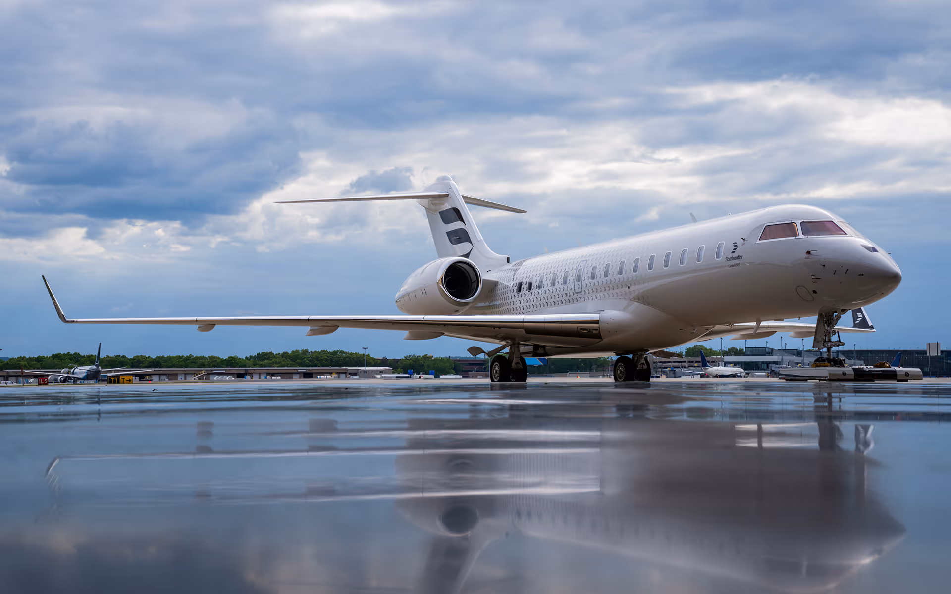 Private jet parked on a wet airport runway under a cloudy sky with its reflection visible on the surface.
