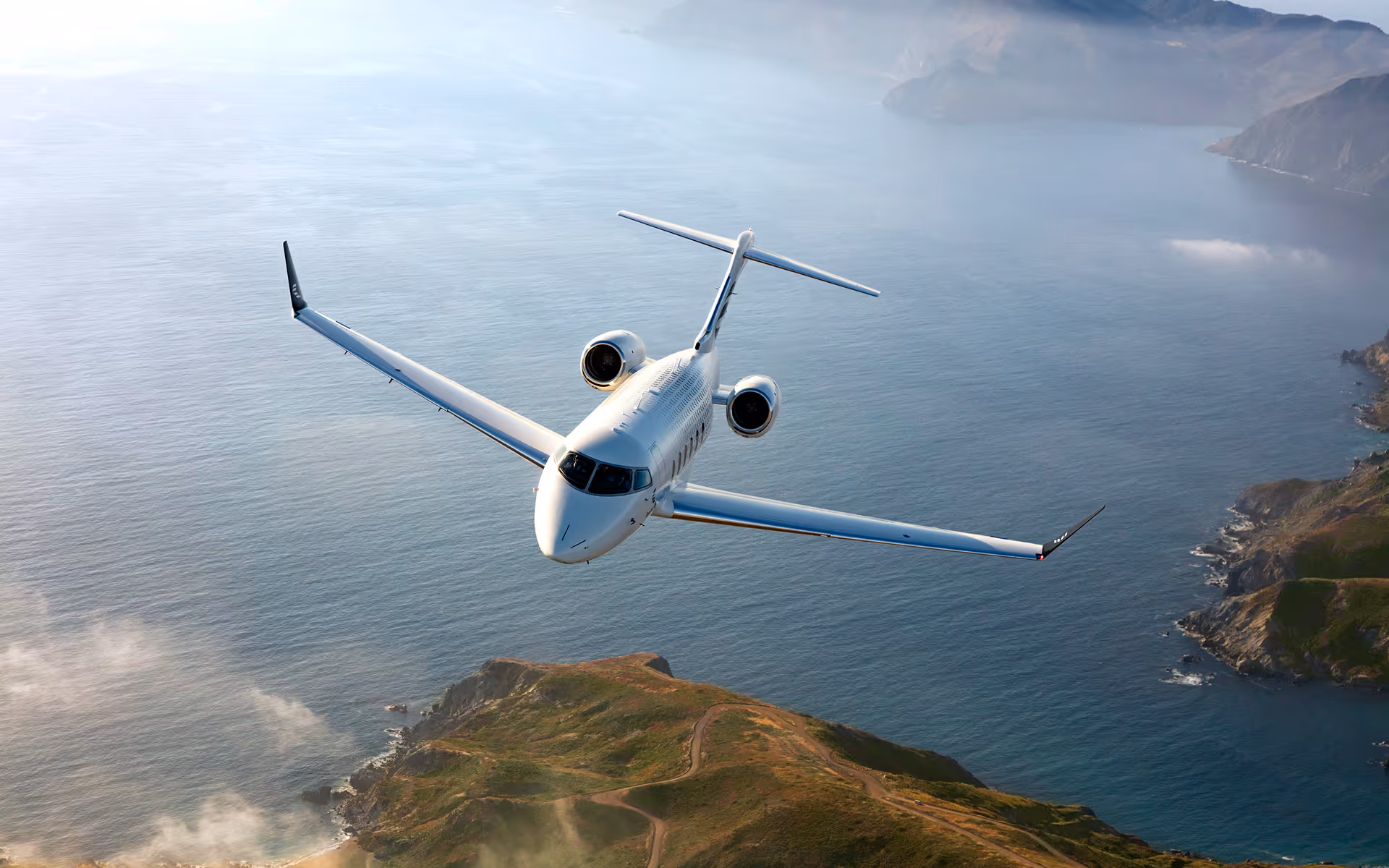 White private jet flying over a coastal landscape with cliffs and ocean beneath.
