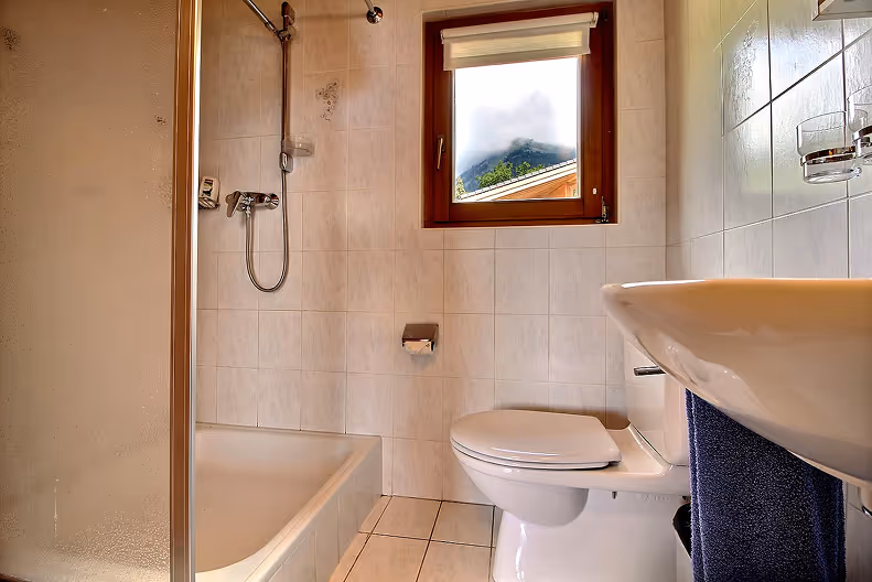 Compact bathroom with a shower, white toilet, white tiled walls, a window showing a foggy mountain, and a white sink with a blue towel.