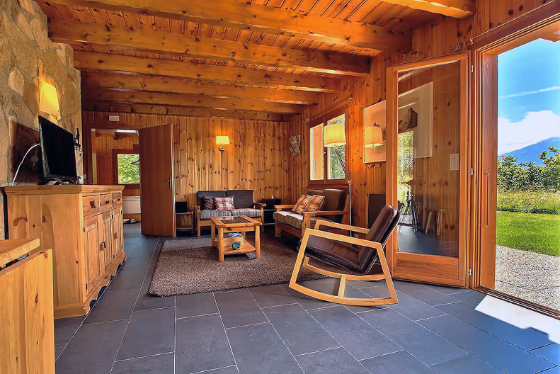 Cozy wooden living room with leather rocking chair, sofas, a TV on wooden cabinet, and open door leading to green lawn and mountain view.