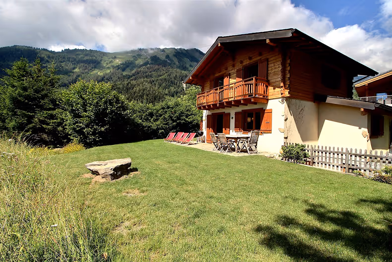 Chalet-style house with wooden balcony, patio furniture, and red lounge chairs on a grassy lawn with forested mountains in the background under a partly cloudy sky.