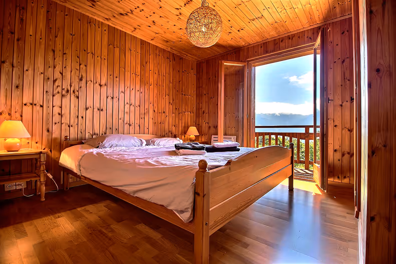 Cozy wooden bedroom with a double bed, two bedside lamps, and an open balcony door showing a view of a sunny sky and greenery.