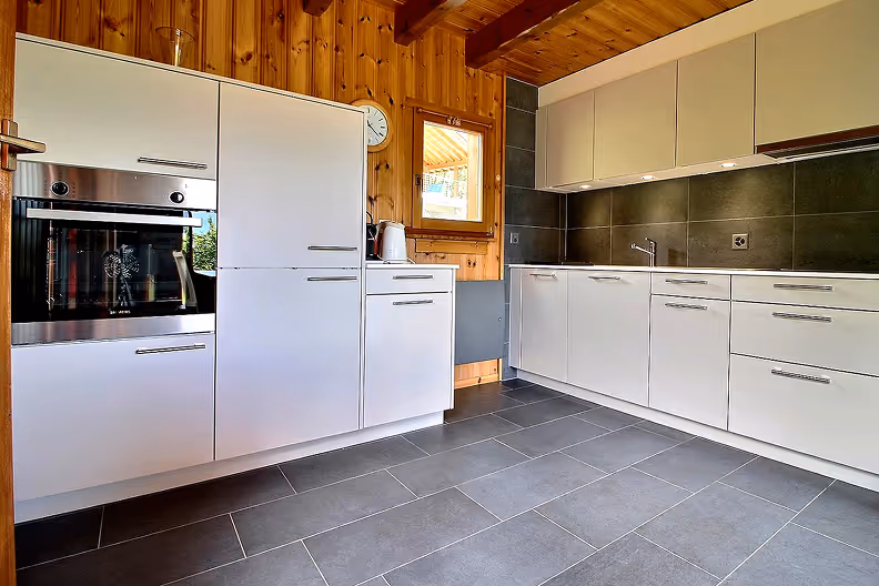 Modern kitchen with white cabinets, stainless steel oven, dark gray tiled floor, wood-paneled walls, and a small window.