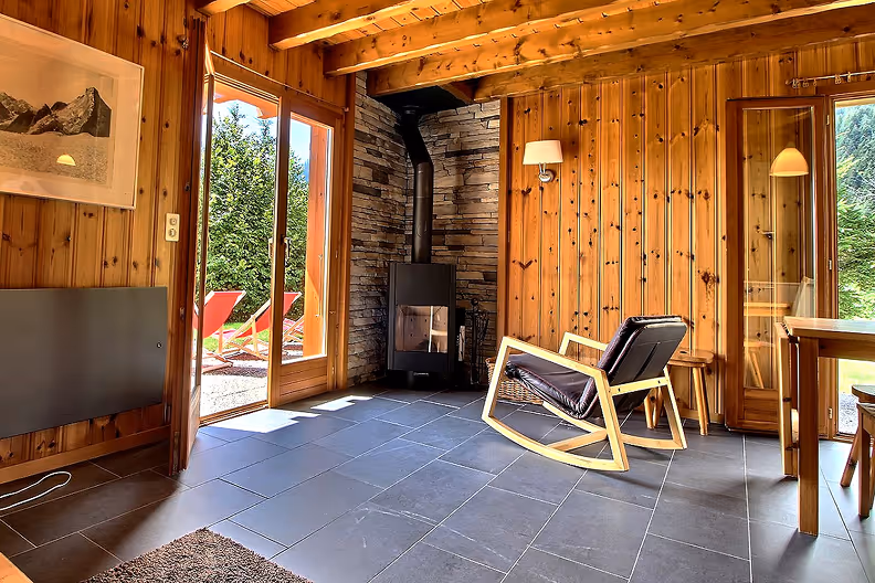 Cozy wooden interior with a black rocking chair, wood stove, and glass doors opening to outdoor red lounge chairs.