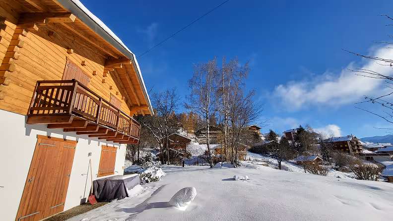 Snow-covered yard beside a wooden chalet with boarded windows and a small balcony under a clear blue sky.