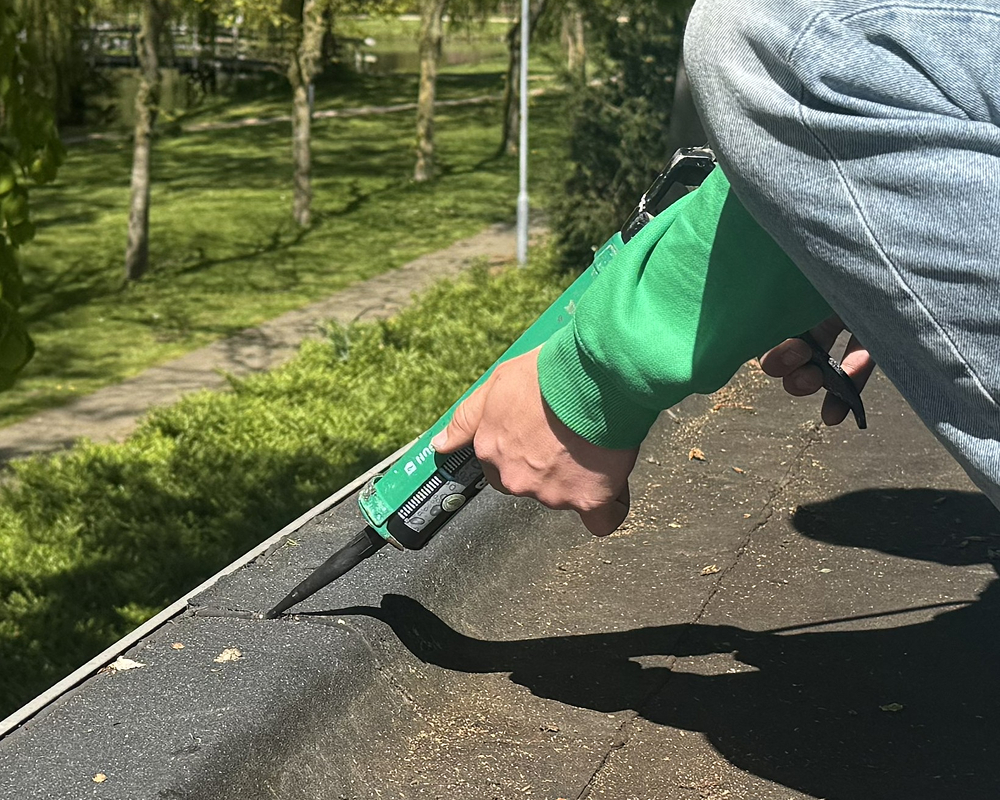 Hand met handschoen die zwarte waterdichte kit aanbrengt rond de dakkoepelflens op een plat dak.