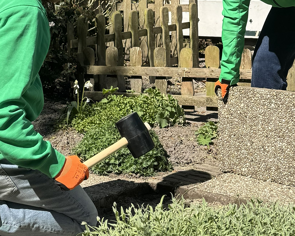 Werknemer met gele handschoenen en zwarte schoenen legt grijze bestratingsstenen op zand, met een rubberen hamer en rolmaat ernaast.